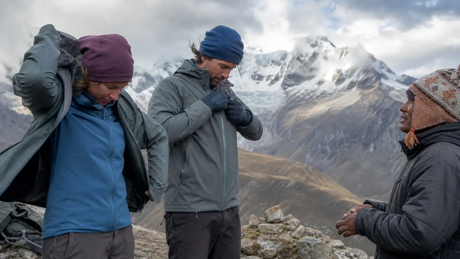 Hikers adding waterproof shells over warm layers with a local guide on a windy Huayhuash mountain pass